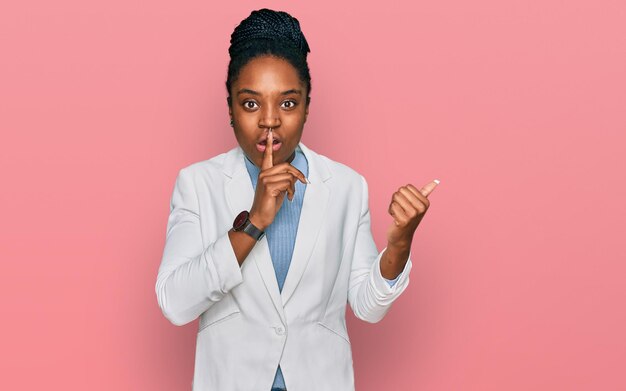 Giovane donna afroamericana che indossa abiti da lavoro chiedendo di stare zitta con il dito sulle labbra che punta con la mano verso il silenzio laterale e il concetto segreto