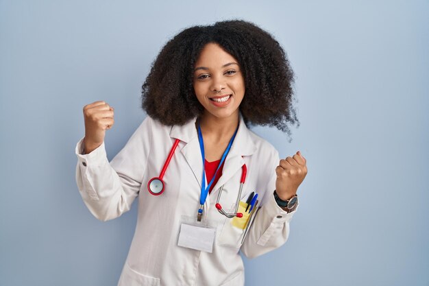 Giovane donna afroamericana che indossa l'uniforme del medico e lo stetoscopio molto felice ed eccitato facendo il gesto del vincitore con le braccia alzate sorridendo e urlando per il concetto di celebrazione del successo