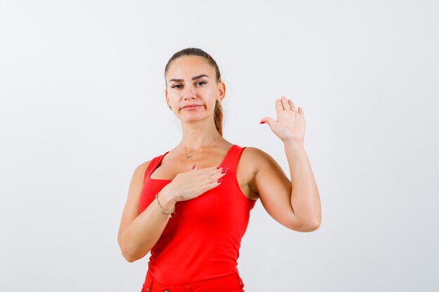 Giovane donna agitando la mano per il saluto in canottiera rossa, pantaloni e guardando fiducioso. vista frontale.