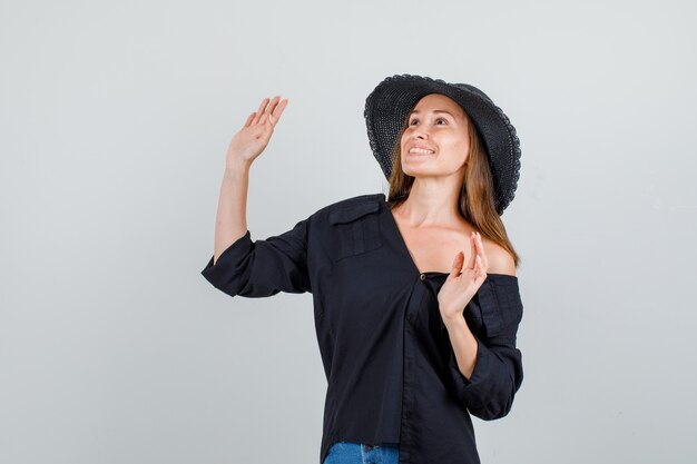 Giovane donna agitando le mani mentre cerca in camicia, pantaloncini, cappello e sembra allegra