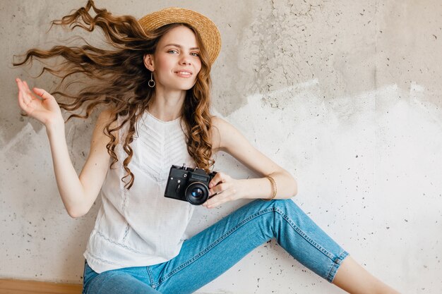 Giovane donna alla moda abbastanza sorridente che indossa jeans blu denim e camicia bianca contro il muro in cappello di paglia