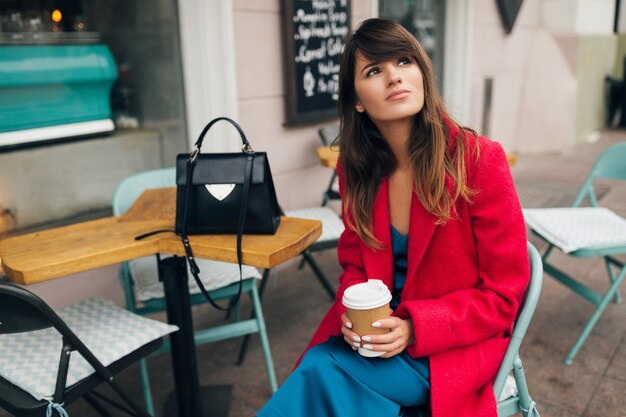 Giovane donna alla moda attraente che si siede nel caffè della via della città nel caffè bevente del cappotto rosso che porta vestito blu