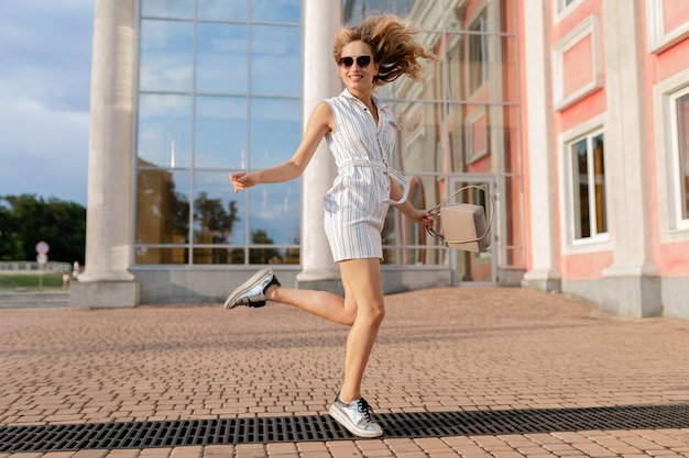 Giovane donna alla moda attraente in esecuzione saltando divertente in scarpe da ginnastica in strada cittadina in abito bianco stile moda estate indossando occhiali da sole e borsa