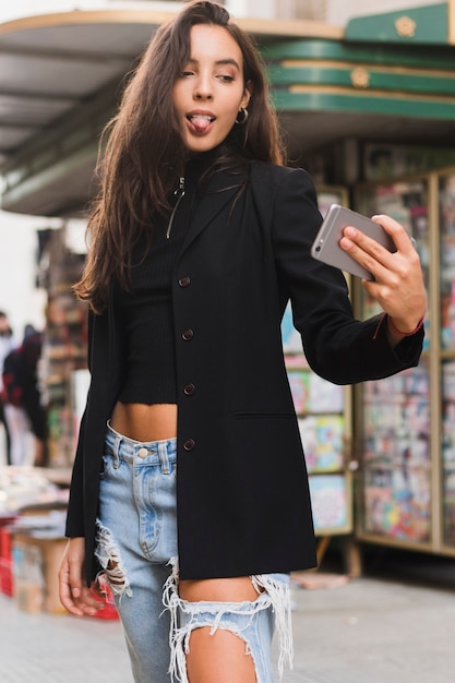 Giovane donna alla moda che attacca la sua lingua fuori mentre prendendo selfie sul telefono cellulare