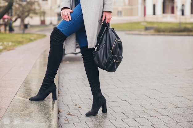 Giovane donna alla moda che cammina nella città d'autunno, stagione fredda, indossa stivali neri con i tacchi alti, zaino in pelle, accessori, cappotto grigio, seduto sulla panchina, tendenza della moda, dettagli del primo piano delle gambe