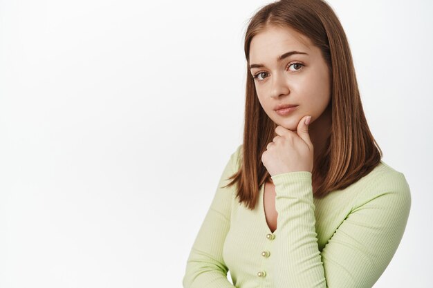 Giovane donna alla moda che pensa, guarda premurosa davanti, sorride e tocca il mento, medita con smth, ascolta con interesse, in piedi contro il muro bianco.