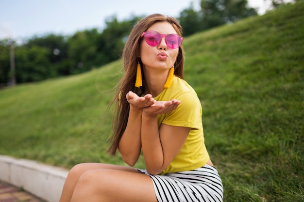 Giovane donna alla moda che ride divertendosi nel parco cittadino, sorridente umore allegro, indossa un top giallo, minigonna a righe, occhiali da sole rosa, tendenza della moda in stile estivo
