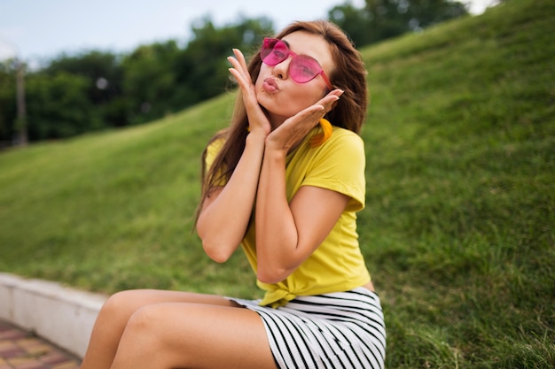 Giovane donna alla moda che ride divertendosi nel parco cittadino, sorridente umore allegro, indossa un top giallo, minigonna a righe, occhiali da sole rosa, tendenza della moda in stile estivo