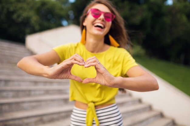 Giovane donna alla moda che ride divertendosi nel parco cittadino, sorridente umore allegro, indossa un top giallo, occhiali da sole rosa, tendenza moda stile estivo, mostrando il segno del cuore