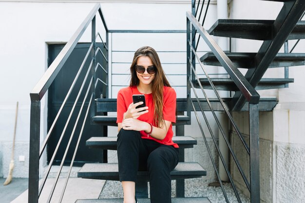 Giovane donna alla moda che si siede sulla scala usando smartphone