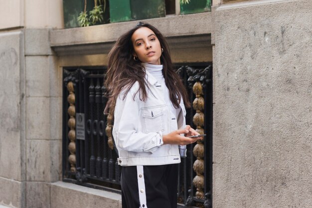 Giovane donna alla moda che tiene in mano il telefono cellulare che guarda l&#39;obbiettivo