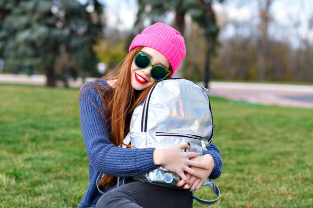 Giovane donna alla moda hipster che si diverte all'aperto, indossa un maglione accogliente con fieno e occhiali da sole, viaggia con lo zaino, emozioni sorprese, stile street city.