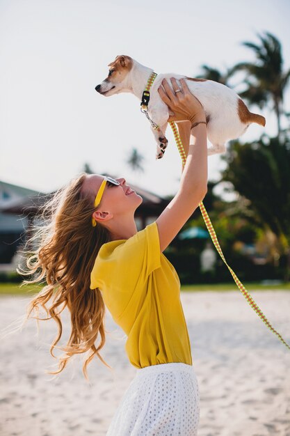 Giovane donna alla moda hipster che tiene camminare e giocare con il cane nel parco tropicale, sorridere e divertirsi