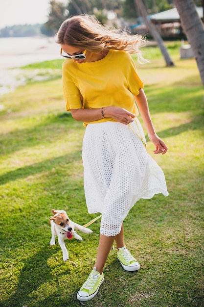 Giovane donna alla moda hipster che tiene camminare giocando cucciolo di cane jack russell, parco tropicale, sorridente e divertirsi, vacanze, occhiali da sole, berretto, camicia gialla, spiaggia di sabbia