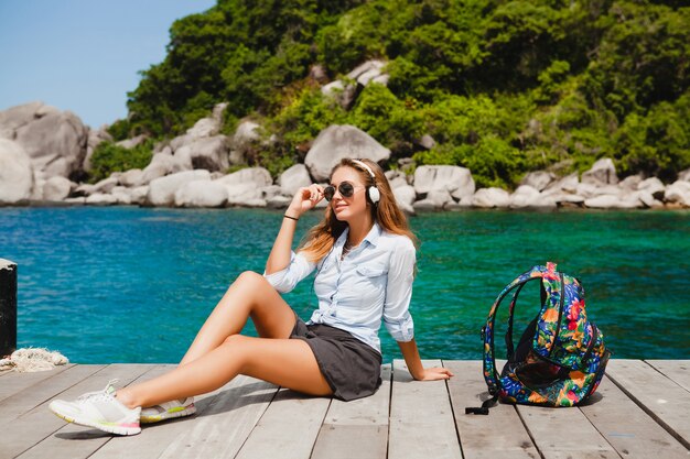 Giovane donna alla moda hipster in viaggio per il mondo, seduta sul molo, occhiali da sole aviator, cuffie, ascolto di musica, vacanza, zaino, camicia di jeans, felice, laguna isola tropicale