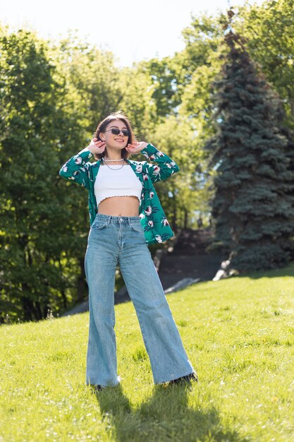 Giovane donna alla moda in camicia verde casuale e jeans al giorno pieno di sole che posa sull'erba nel parco