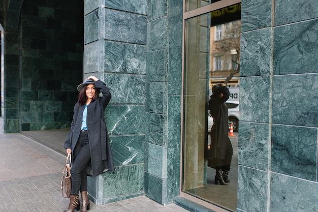 Giovane donna alla moda in cappotto grigio, cappello che cammina sulla strada nel centro della città. Sorridente, emozioni vere, stile di vita elegante, vestiti di lusso, look elegante.