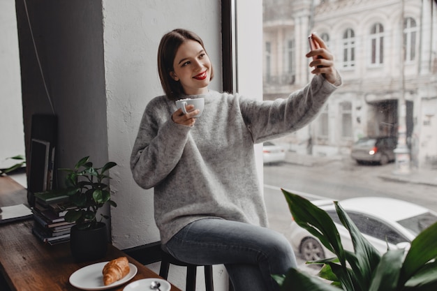 Giovane donna alla moda in maglione grigio e jeans in posa con una tazza di cappuccino e prendendo selfie dalla finestra.