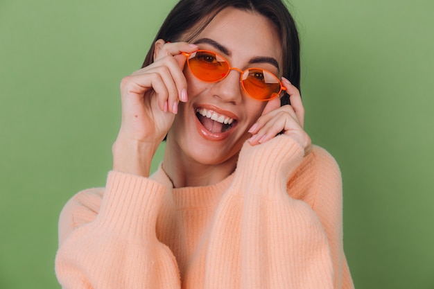Giovane donna alla moda in maglione pesca casual e vetri arancioni isolati sulla risata sorridente positiva felice della parete verde oliva intorno allo spazio della copia