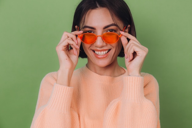 Giovane donna alla moda in maglione pesca casual e vetri arancioni isolati sulla risata sorridente positiva felice della parete verde oliva intorno allo spazio della copia
