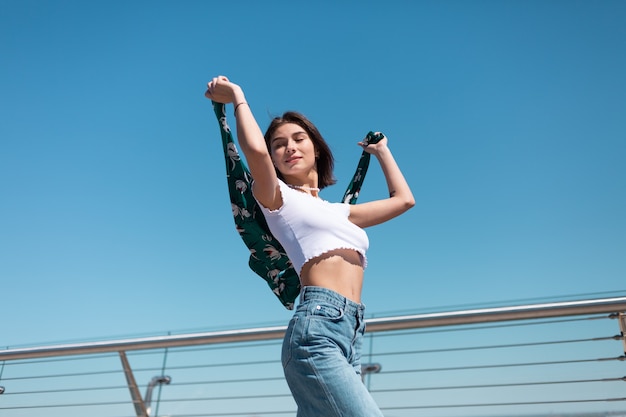 Giovane donna alla moda in top corto casual bianco e jeans in posa sul ponte della città in una giornata calda e soleggiata