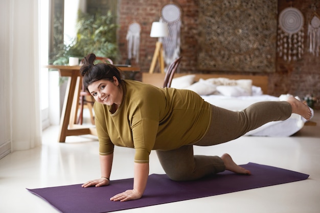 Giovane donna allegra autodeterminata con corpo sinuoso e nodo dei capelli che si esercita all'interno sulla stuoia di yoga che rinforza i muscoli, mantenendo entrambe le mani e il ginocchio sul pavimento, sollevando una gamba e sorridendo gioiosamente