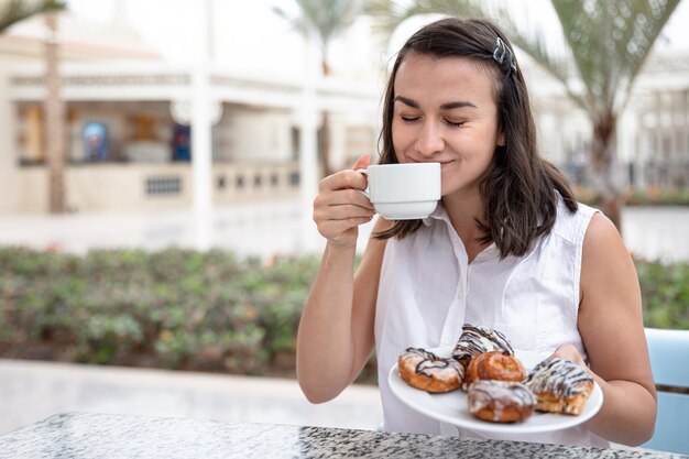 Giovane donna allegra che gode del caffè del mattino con ciambelle sulla terrazza all'aperto. Concetto di vacanza e ricreazione.