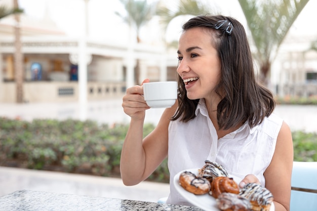 Giovane donna allegra che gode del caffè del mattino con ciambelle sulla terrazza all'aperto
