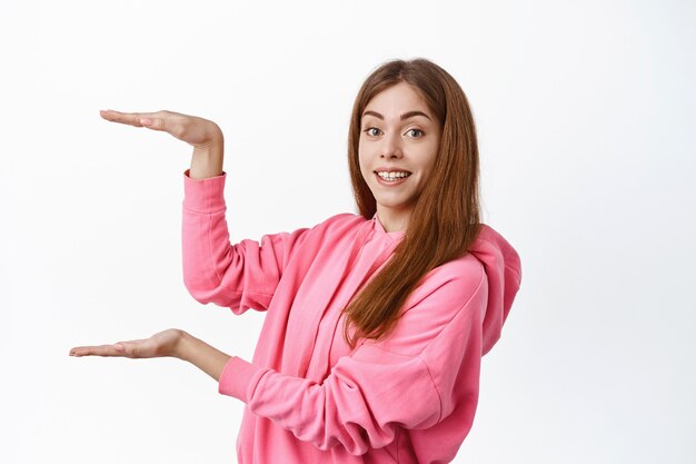 Giovane donna allegra che mostra grandi dimensioni su copyspace, display prodotto o logo, tenendo lo spazio vuoto per banner, in piedi sul muro bianco