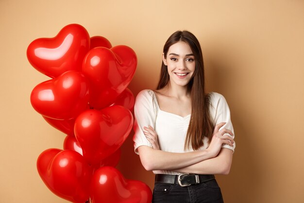 Giovane donna allegra che sembra felice il giorno di San Valentino in piedi vicino a palloncini di cuori con le braccia incrociate...