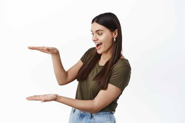 Giovane donna allegra che tiene il tuo logo in mano e sorridente Ragazza eccitata che fa un gesto di casella vuota sullo spazio della copia come se tenesse qualcosa di sfondo bianco