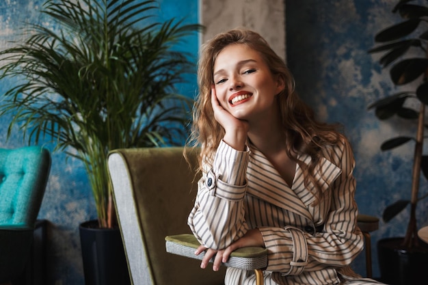 Giovane donna allegra con capelli mossi in trench a righe appoggiato a mano felicemente guardando nella fotocamera mentre si trascorre del tempo in un caffè moderno
