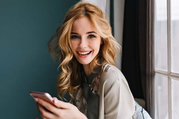 Giovane donna allegra con lunghi capelli ondulati con smartphone rosa nelle mani alla ricerca e sorridente, seduto accanto alla finestra. Indossare un pigiama di seta. Muro turchese, tende.