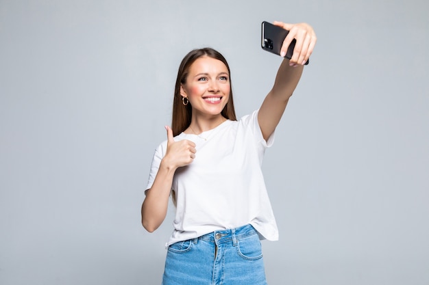 Giovane donna allegra felice che mostra pollice in su e che fa selfie sullo smartphone isolato