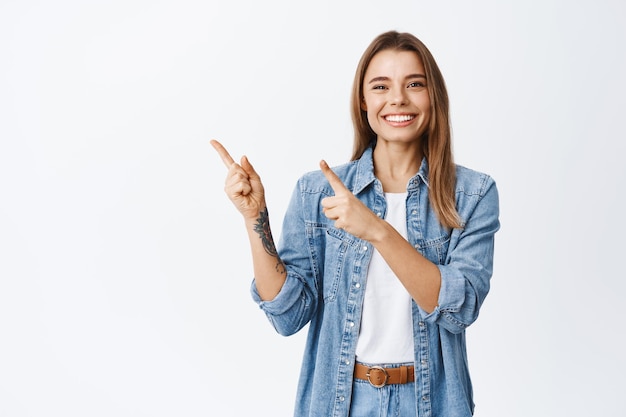 Giovane donna allegra in abiti casual che mostra pubblicità, puntando il dito nell'angolo in alto a sinistra e sorridendo, consigliando l'offerta, in piedi su bianco