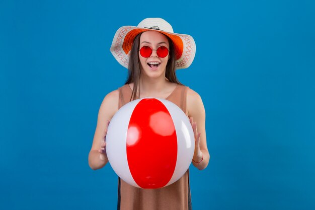 Giovane donna allegra in cappello estivo che indossa occhiali da sole rossi tenendo palla gonfiabile con il sorriso sul viso in piedi sopra lo spazio blu