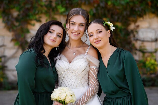 Giovane donna allegra sposa elegante in posa con due damigelle d'onore in abito verde scuro in piedi all'aperto Donna attraente in abito da sposa con maniche lunghe Celebrazione del matrimonio Amici della sposa