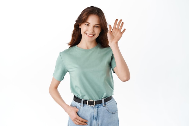 Giovane donna amichevole agitando la mano e salutando, salutando gli ospiti, facendo un gesto e sorridendo ampiamente, in piedi su sfondo bianco