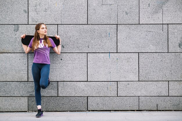 Giovane donna appoggiata a un muro urbano piastrellato grigio in piedi su una gamba