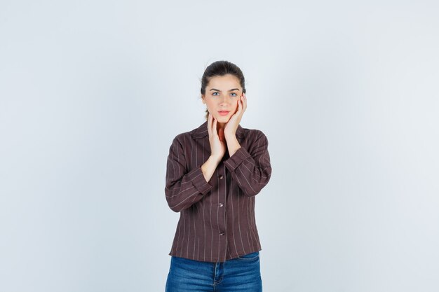 Giovane donna appoggiata guancia sul palmo, con la mano sulla guancia in camicia a righe, jeans e guardando affascinante, vista frontale.