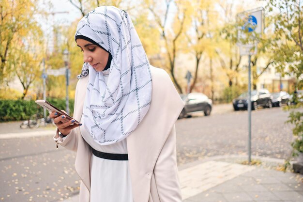 Giovane donna araba attraente in hijab che utilizza intensamente lo smartphone sull'accogliente strada della città