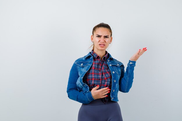 Giovane donna arrabbiata in camicia a scacchi, giacca, pantaloni e sguardo esitante, vista frontale.