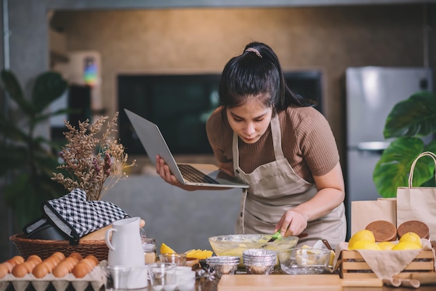 Giovane donna asiatica adulta che prepara prodotti da forno fatti in casa in una cucina a casa guardando sul computer portatile per i metodi di cottura