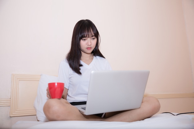 Giovane donna asiatica attraente che per mezzo del computer portatile mentre trovandosi sul letto in abbigliamento casual