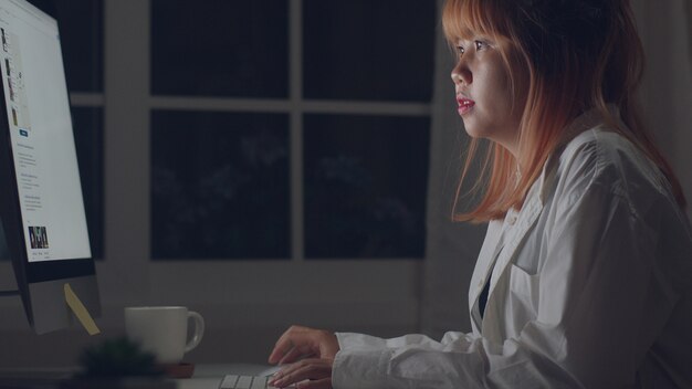 Giovane donna asiatica che lavora tardi usando il desktop sullo scrittorio nel salone a casa.