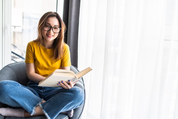 Giovane donna asiatica che legge un libro mentre è seduta in poltrona moderna a casa