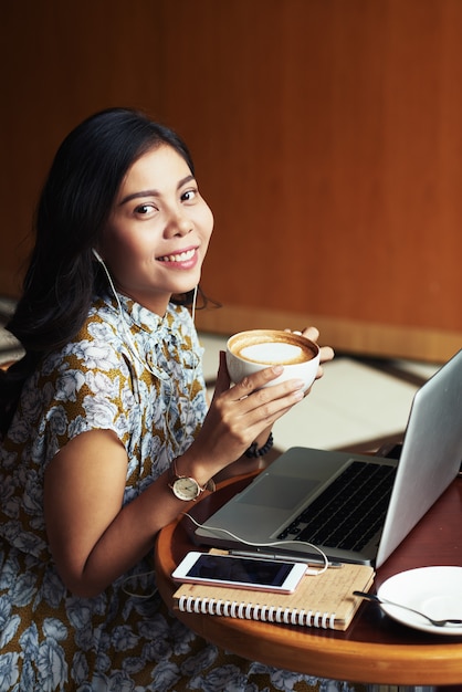 Giovane donna asiatica che si siede con il computer portatile in caffè e che gode del cappuccino