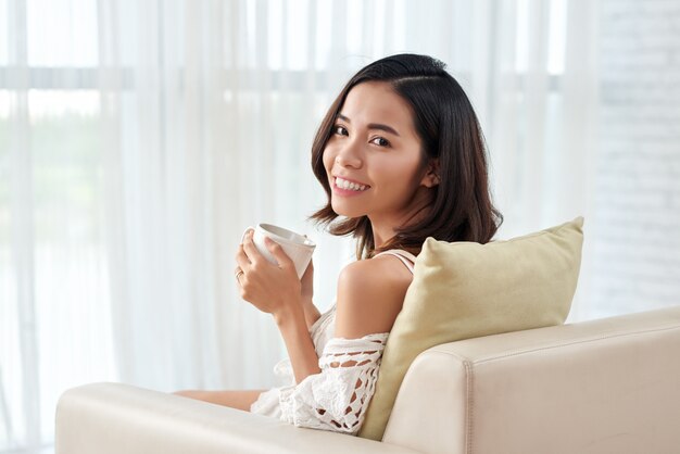 Giovane donna asiatica che si siede in poltrona con la tazza di caffè che esamina macchina fotografica