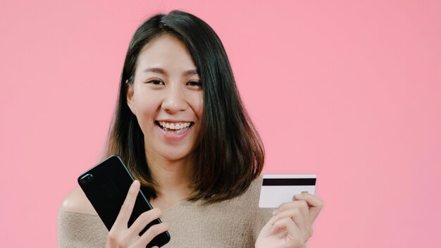 Giovane donna asiatica che utilizza smartphone che compra acquisto online dalla carta di credito che ritiene sorridere felice in abbigliamento casuale sopra il colpo rosa dello studio del fondo.