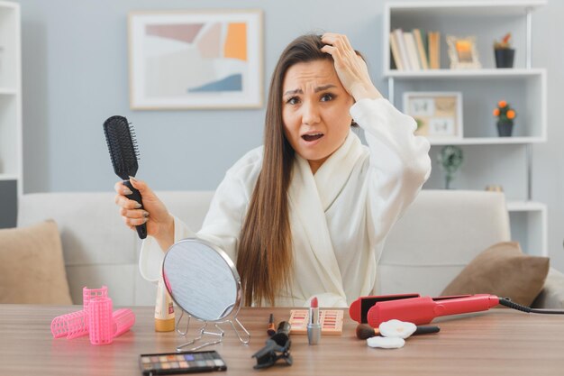 Giovane donna asiatica con i capelli lunghi scuri che si siede al tavolo da toeletta a casa facendo la routine di trucco mattutina che sembra confusa mentre si spazzola i capelli che soffrono di caduta dei capelli